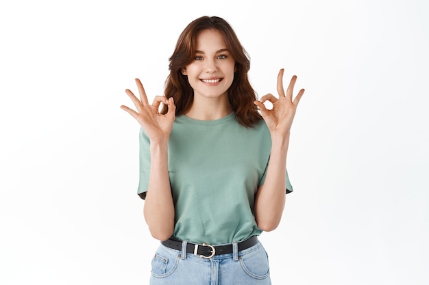 Super bien. Sonriente niña satisfecha alabando y felicitando la elección, mostrando buenos gestos y asintiendo con la cabeza en señal de aprobación, garantía de calidad, recomendando, de pie sobre fondo blanco.