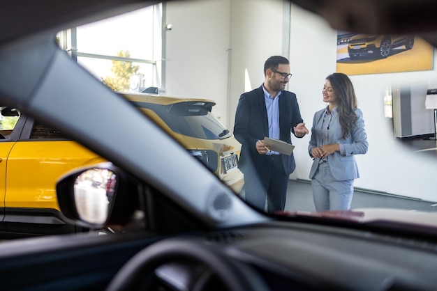 Súper equipo de ventas en el concesionario dos consultores o gerentes en trajes elegantes con computadora portátil y tableta en las armas en el concessionario de automóviles