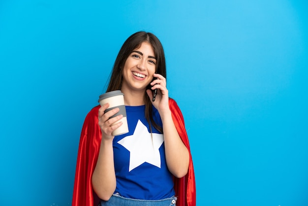 Super Hero kaukasische Frau isoliert auf blauem Hintergrund mit Kaffee zum Mitnehmen und einem Handy