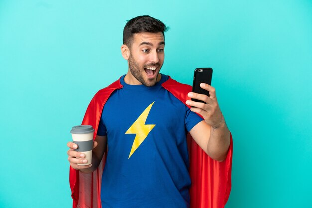 Super Hero kaukasischer Mann isoliert auf blauem Hintergrund mit Kaffee zum Mitnehmen und einem Handy