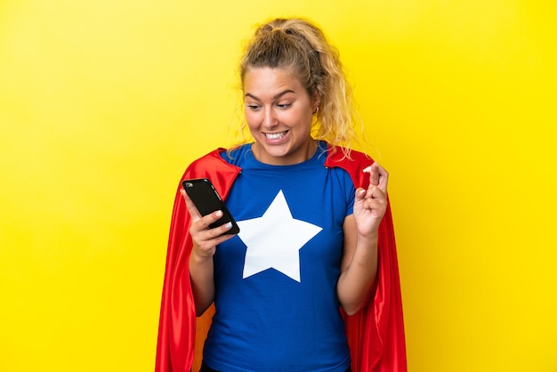 Super Hero mujer aislada sobre fondo amarillo a través de teléfono móvil con los dedos cruzados