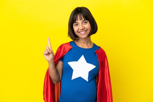 Foto super heroína grávida isolada em fundo amarelo mostrando e levantando um dedo em sinal dos melhores