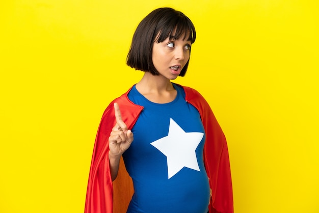 Super-heroína grávida isolada em um fundo amarelo pensando uma ideia apontando o dedo para cima