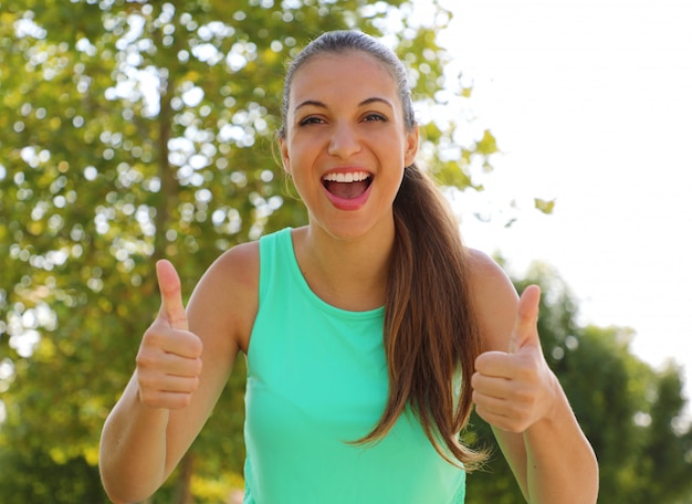 ¡Super mujer! Retrato de niña ganadora mostrando los pulgares para arriba. Mujer sana fitness sonriente positiva al aire libre.