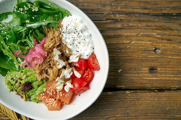 Superalimento - uma tigela com arroz, salmão, guacamole, ovo escalfado e tomate cereja temperado com iogurte grego em uma madeira. alimento equilibrado