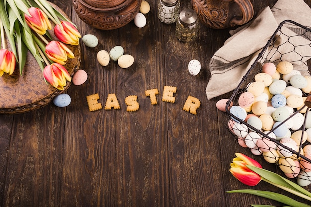 Superfície de Páscoa feliz, letras de biscoitos, tulipas e ovos de codorna coloridos em uma cesta de metal na superfície de madeira