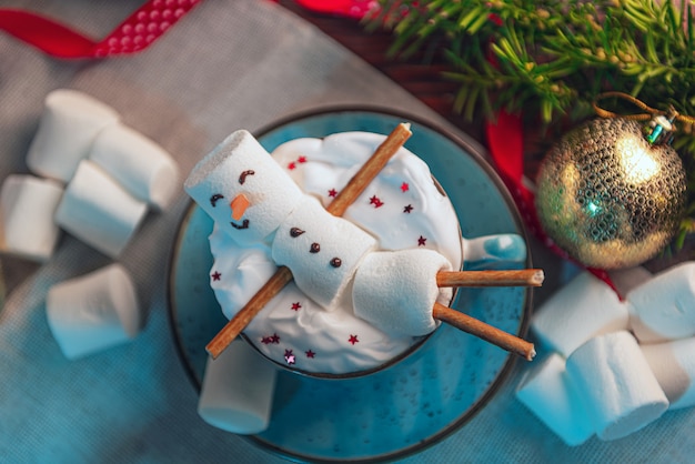 Superficie navideña con un hombre de malvavisco acostado en una taza con cacao, en la superficie un árbol de Navidad con juguetes, una guirnalda y regalos