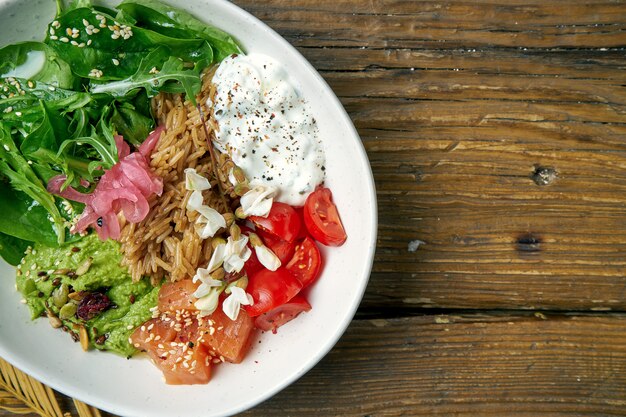 Foto superfood - eine schüssel mit reis, lachs, guacamole, pochiertem ei und kirschtomaten, gewürzt mit griechischem joghurt auf holz. ausgewogenes essen