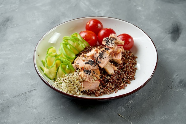 Superfood - Quinoa mit gebackenem Lachs, Gurke und Tomaten in einer weißen Schüssel auf einem grauen Tisch. Nahaufnahme, selektiver Fokus