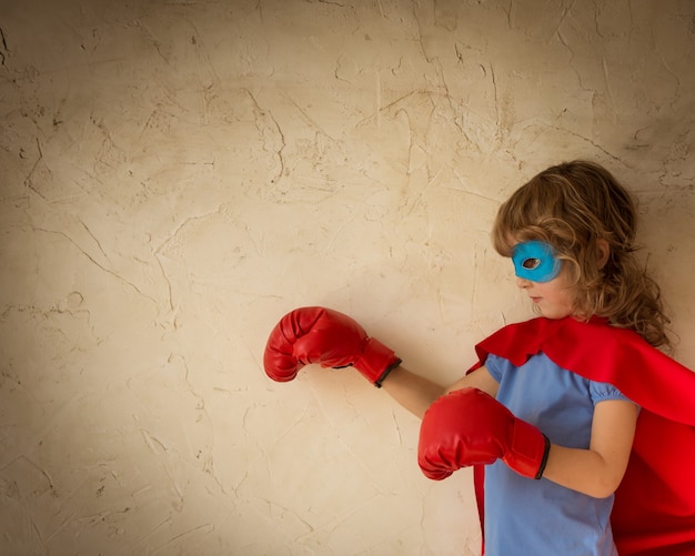 Superheldenkind in rotem Umhang, Boxhandschuhen und blauer Maske vor Grunge-Wandhintergrund