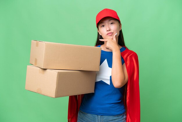 Superhéroe entrega mujer asiática sosteniendo cajas sobre fondo aislado pensando