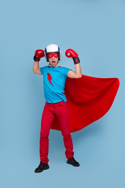 Superhéroe masculino fuerte valiente en traje y guantes de boxeo que muestra el poder sobre fondo azul en el estudio y mirando a la cámara