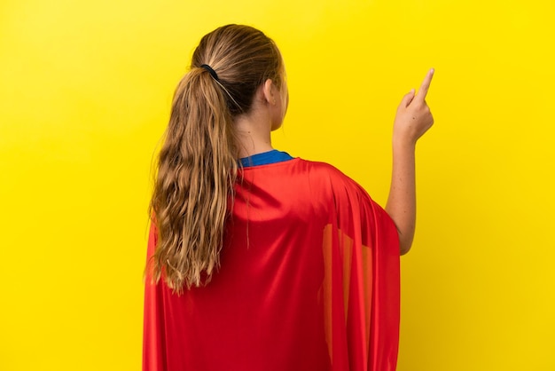 Superhéroe niña aislada sobre fondo amarillo apuntando hacia atrás con el dedo índice