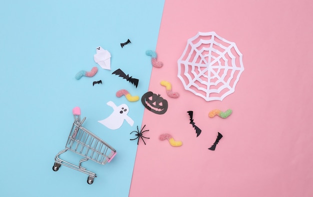 Supermarktwagen mit handgemachtem Halloween-Papierdekor, gummiartige Würmer auf rosa blauem Pastellhintergrund. Ansicht von oben