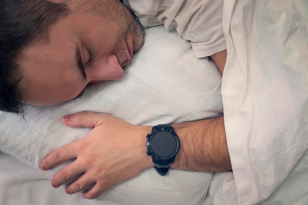 Supervisión de los latidos del corazón con un dispositivo inteligente en un hombre de ensueño duerme sobre una almohada blanca con un reloj inteligente en la muñeca para controlar la pantalla en blanco de los latidos del corazón en la pantalla del reloj inteligente en blanco