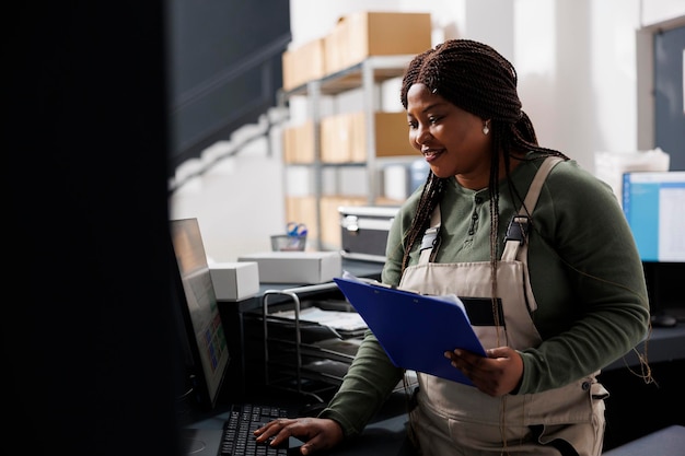 Supervisor de almacén comprobando los pedidos en línea de los clientes en la computadora antes de comenzar a preparar los paquetes para su envío en el almacén Trabajador afroamericano con un mono industrial de pie en el mostrador