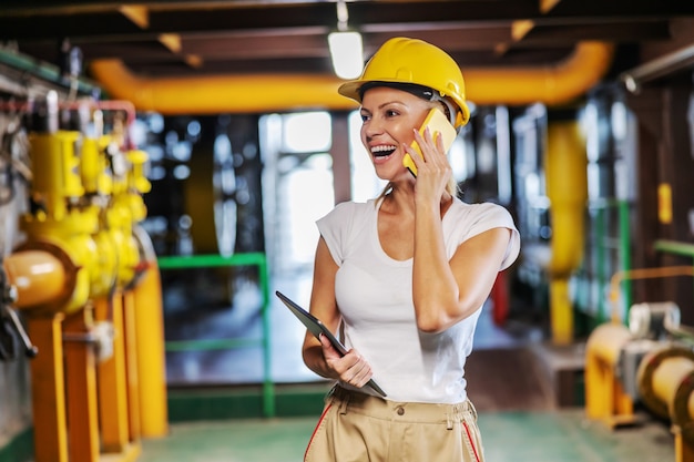 Supervisor feminino sorridente de meia idade em uniforme de trabalho com capacete na cabeça, segurando o tablet e tendo uma conversa telefônica em pé na planta de aquecimento.