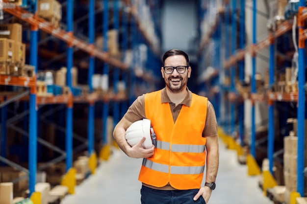 Un supervisor orgulloso posando en el almacenamiento con casco en las manos y sonriendo a la cámara