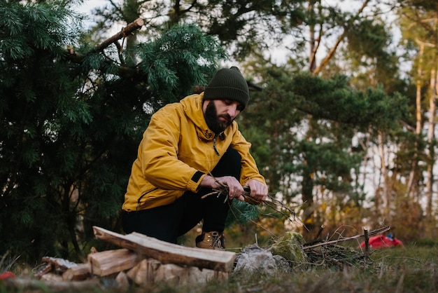 Supervivencia en la naturaleza Un hombre barbudo enciende una fogata cerca de un refugio improvisado hecho con ramas de pino