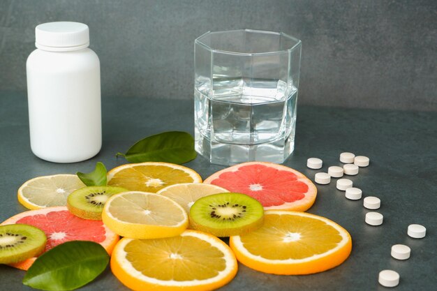 Suplementos ou alimentos naturais saudáveis Pílulas brancas espalhadas vitaminas Frutas cítricas cortadas laranja limão kiwi uvas copo com água em um fundo cinza pedra