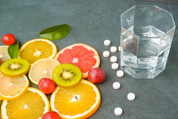 Suplementos ou alimentos naturais saudáveis Pílulas brancas espalhadas vitaminas Frutas cítricas cortadas laranja limão kiwi uvas copo com água em um fundo cinza pedra