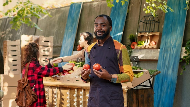 Suporte de estande masculino com avental segurando produtos biológicos maduros para mostrar produtos de agricultura orgânica aos clientes. Agricultor de homem alegre vendendo frutas e legumes caseiros ao ar livre. Tiro portátil.