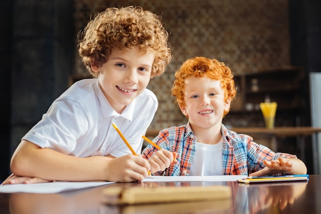 Suporte é o que realmente importa. Irmãos amigáveis, gostando de passar tempo juntos e sorrindo amplamente para a câmera enquanto desenham com lápis coloridos.