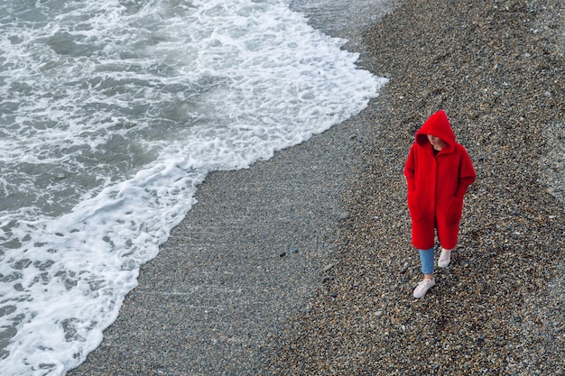 Surf de mar, orilla de guijarros. Una mujer con abrigo rojo con capucha camina por la playa, día de invierno.