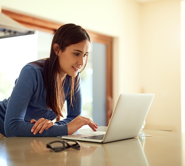 Surf en el mostrador de la cocina Foto de una mujer joven feliz usando su computadora portátil mientras está de pie en su cocina