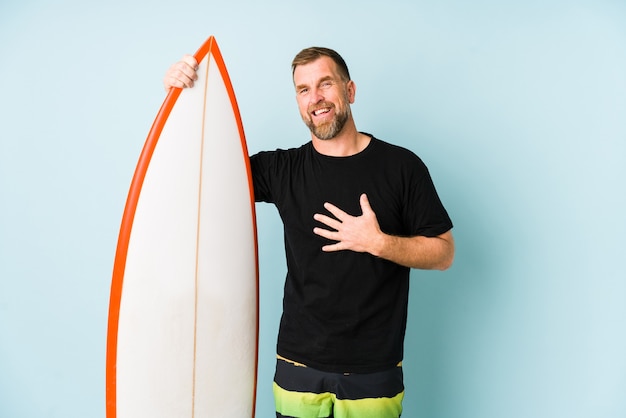 Surfen hombre aislado sobre fondo azul se ríe a carcajadas manteniendo la mano en el pecho.