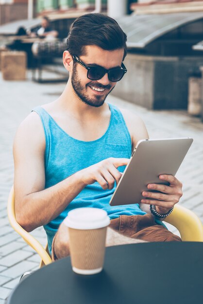 Surfen im Internet vom Café. Hübscher junger Mann in Freizeitkleidung, der ein digitales Tablet hält und es mit einem Lächeln betrachtet, während er im Straßencafé im Freien sitzt