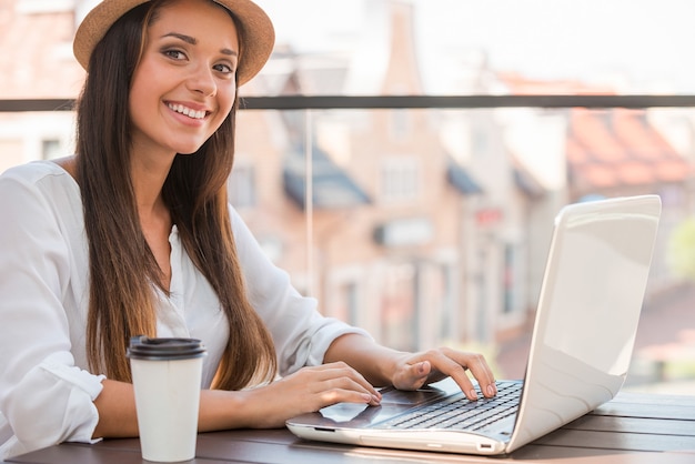 Surfen im Netz im Freien. Schöne junge Frau mit flippigem Hut, die am Laptop arbeitet und lächelt, während sie draußen sitzt