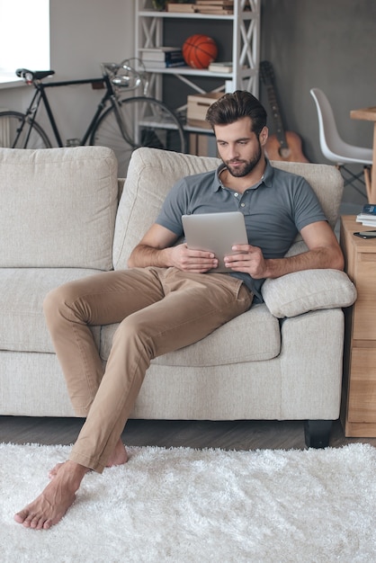 Surfen im Netz zu Hause. Hübscher junger Mann, der sein digitales Tablet benutzt, während er zu Hause auf der Couch sitzt