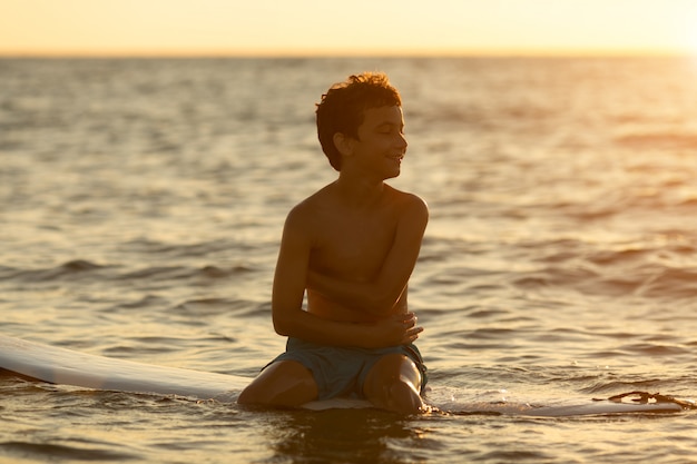Surfer boy sentado en una tabla de surf al amanecer