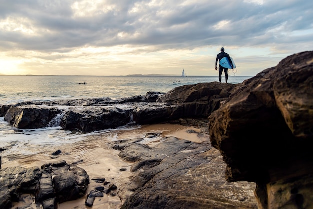 Surfer, der auf Felsen steht und die Wellen beobachtet, bevor er an einem sonnigen Tag bei Sonnenuntergang in Noosa Heads, Queensland, Australien, ins Wasser springt