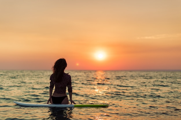 Surfermädchen, das Ozeanstrandsonnenuntergang betrachtend surft