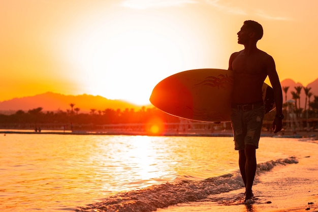 Foto surfista de homem de silhueta na luz do sol andando pela praia com prancha de surf perto da beira da água aproveitando o hobby de prática de horário de verão durante férias de fuga cara recriando na ilha tropical com ondas
