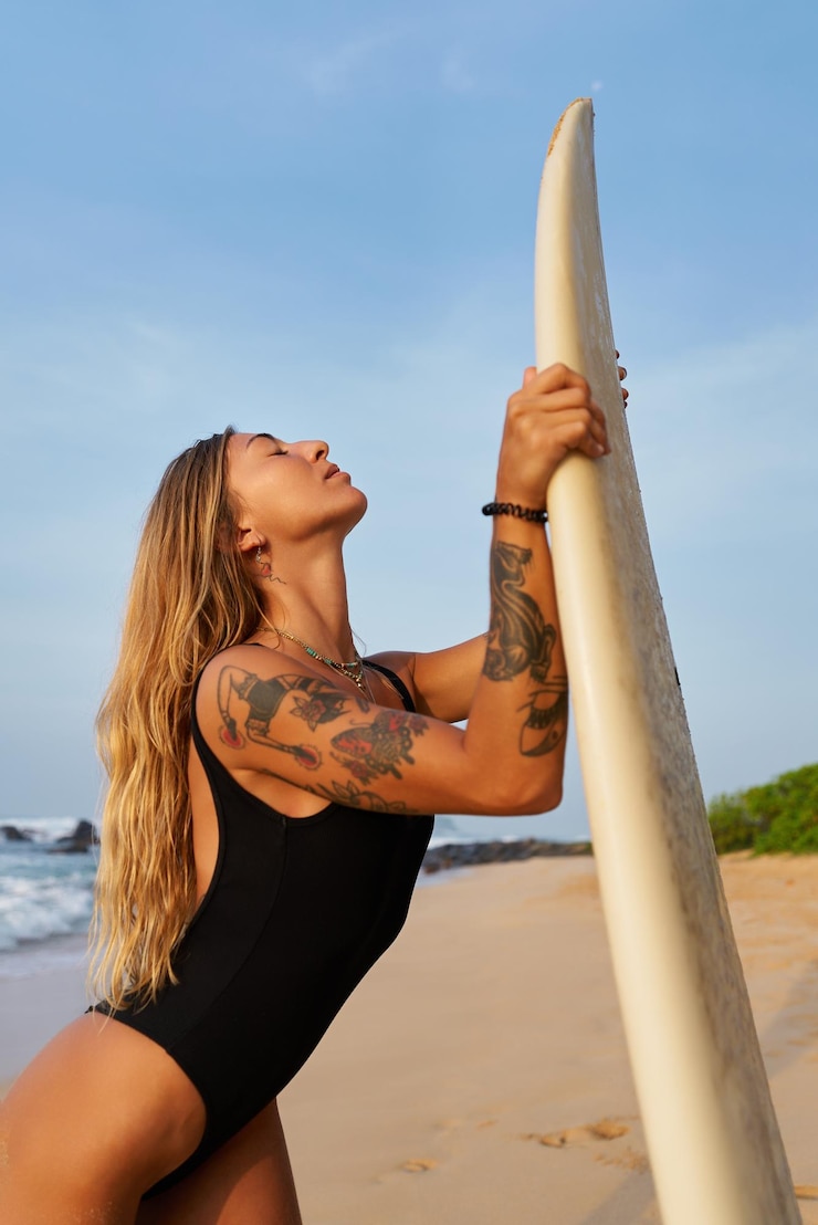 Foto surfista jovem tatuada segura uma prancha de surf em uma praia ensolarada, cabelo loiro, fato de banho, ondas do oceano, fitness.