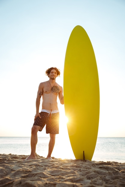 Surfista joven sonriente sosteniendo la tabla de surf mientras está de pie en la playa al atardecer