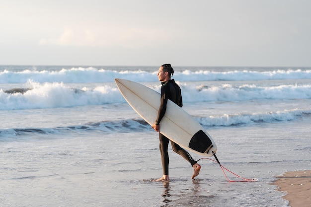 Foto surfista latino caminando y sonriendo en la playa con una tabla de surf metiéndose en el océano