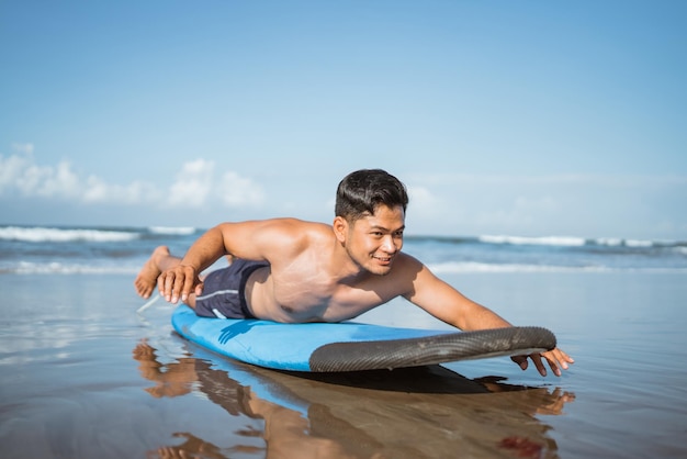Surfista masculino asiático rema en una tabla de surf en la arena antes de entrenar en la playa
