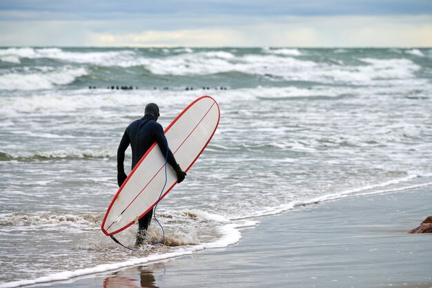 Surfista masculino em maiô preto caminhando à beira-mar e segurando uma prancha de surf branca na mão