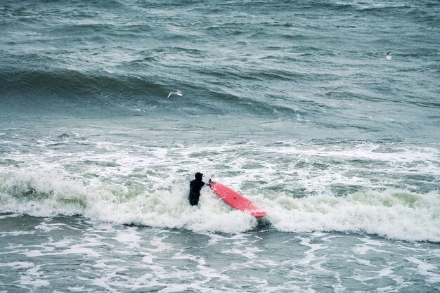 Surfista masculino em maiô preto no oceano com uma prancha de surf vermelha esperando uma grande onda. Dia quente, belas águas do mar, cenário natural. Esporte e conceito de atividade ao ar livre.