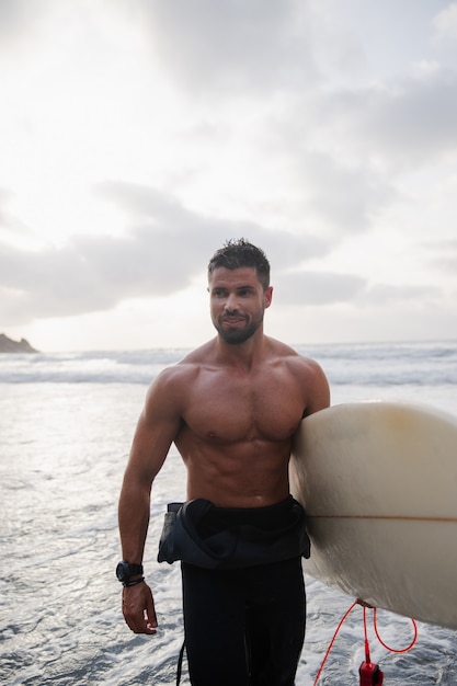 Surfista masculino con tabla de surf en la playa al atardecer, él es joven - Hombre atractivo sonriente divirtiéndose y haciendo deporte extremo - felicidad del concepto de los millennials - foco en su rostro.