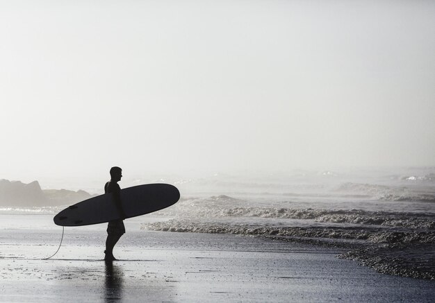 Foto surfista segurando uma prancha de surf enquanto está de pé na costa na praia