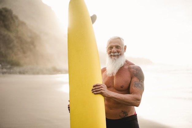 Surfista sênior segurando uma prancha de surf na praia ao pôr do sol - foco no rosto