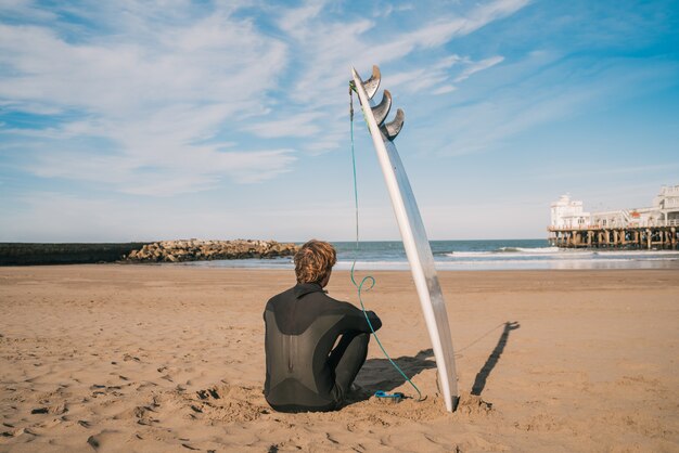 Surfista sentado en la playa de arena y junto a la tabla de surf.