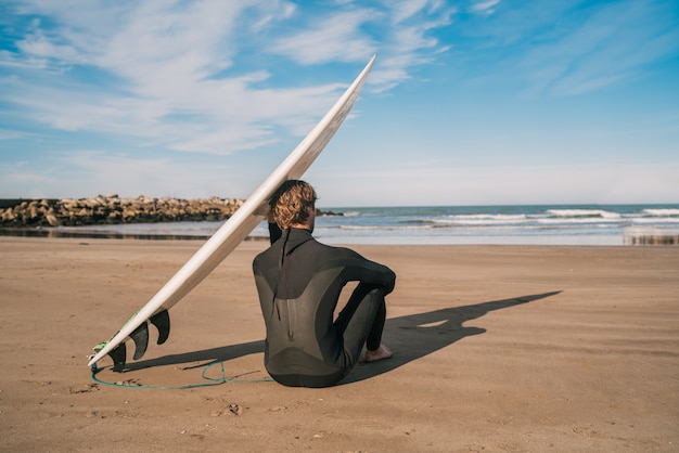 Surfista sentado en la playa de arena y junto a la tabla de surf.