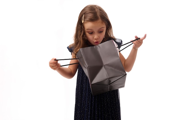 Foto surpreendeu a linda menina de 4 anos menina com um vestido de noite azul escuro, olhando para um pacote de compras preto, isolado no fundo branco, com espaço de cópia. conceito de black friday