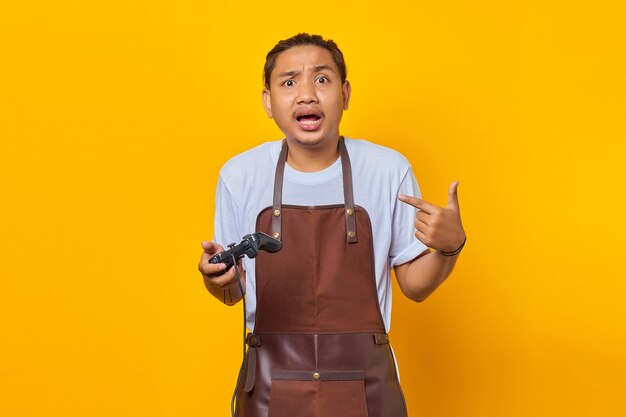 Surpreendeu um jovem asiático bonito usando avental, segurando o controle do jogo e apontando para si mesmo